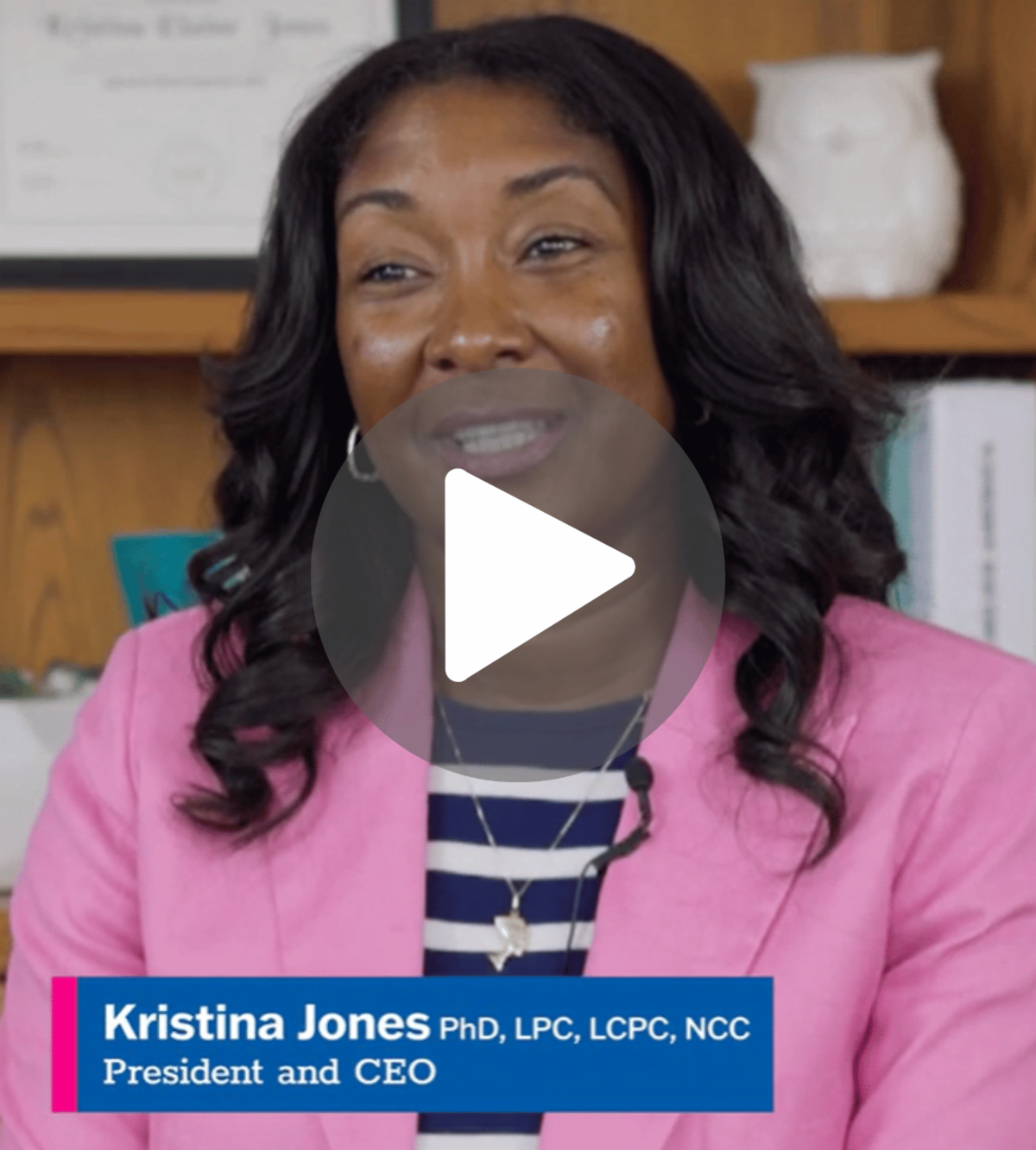 Dr Jones sits in front of a bookshelf wearing a pink blazer over a blue and white striped shirt.