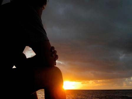 Man looking out on the ocean, dark clouds overhead but the sun shines on the horizon