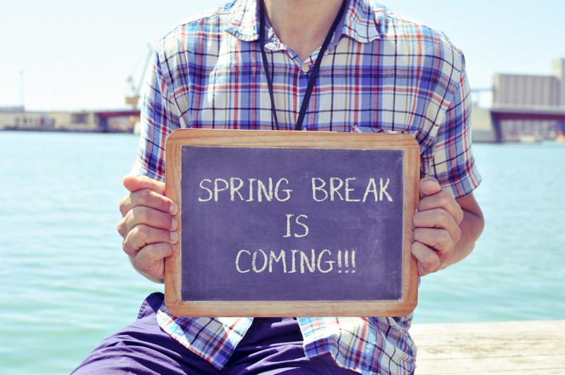 a young caucasian man sitting in a wooden pier shows a chalkboard with the text spring break is coming written in it, with a filter effect