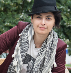 The poet and organizer Rasha Abdulhadi looks directly at the camera. They wear a black fedora and a wine colored blazer and a black and white scarf.